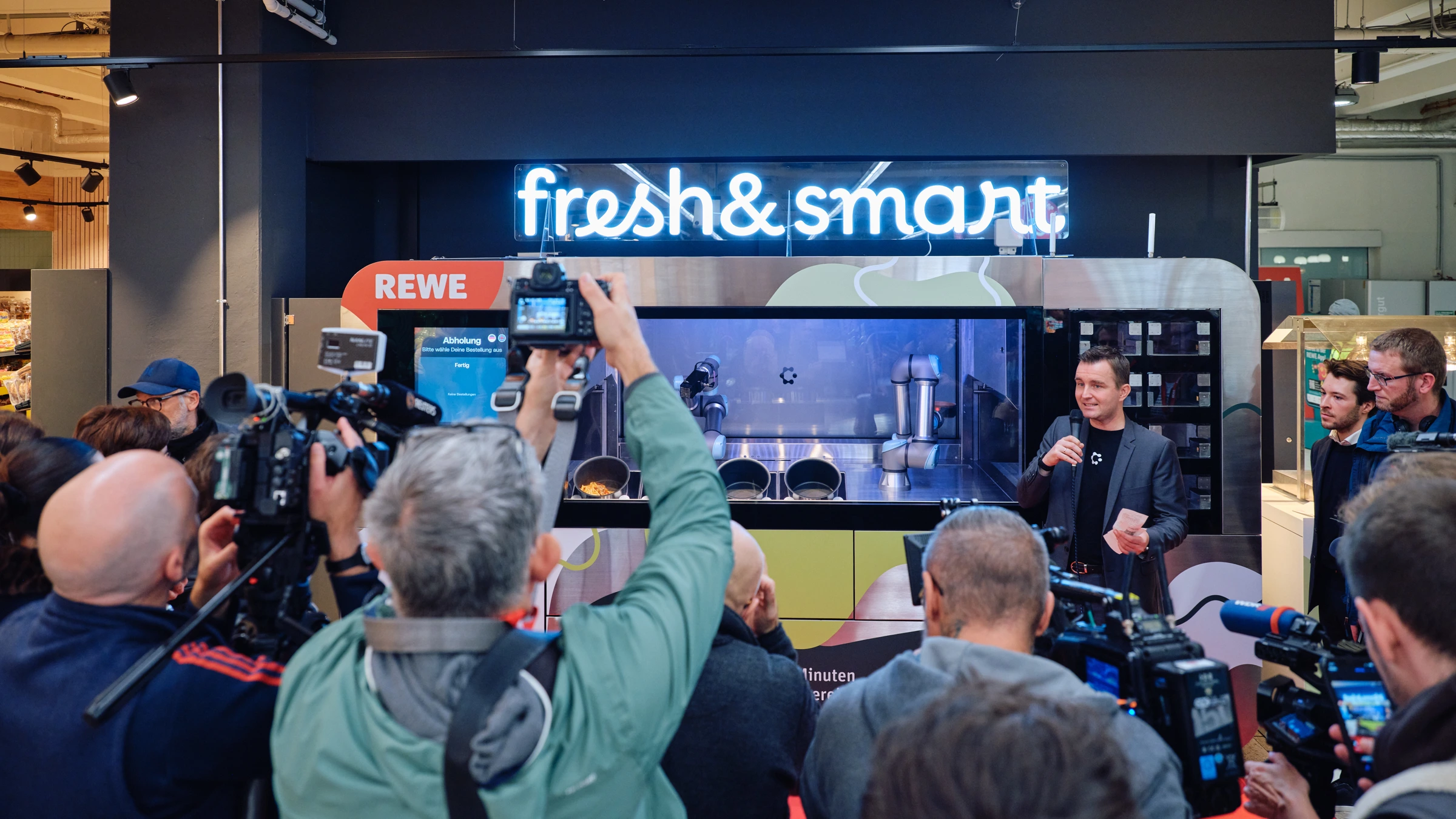 Circus cooking robot behind glass at a REWE Fresh & Smart press event, with journalists in front.