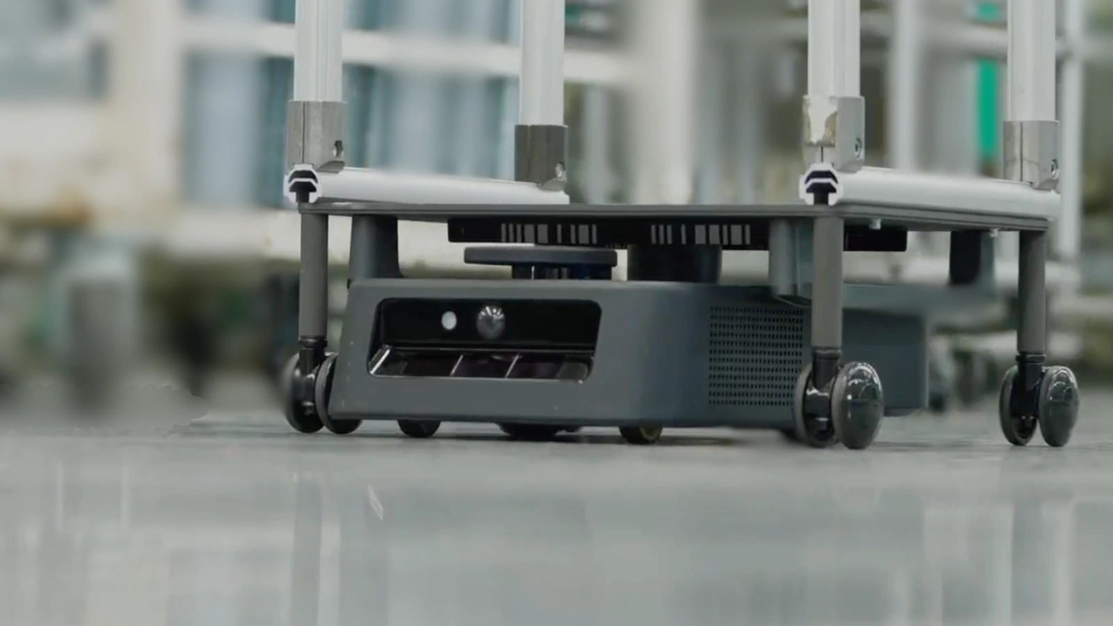 A dark Kachaka-series mobile robot beneath a metal trolley with caster wheels on a factory floor.