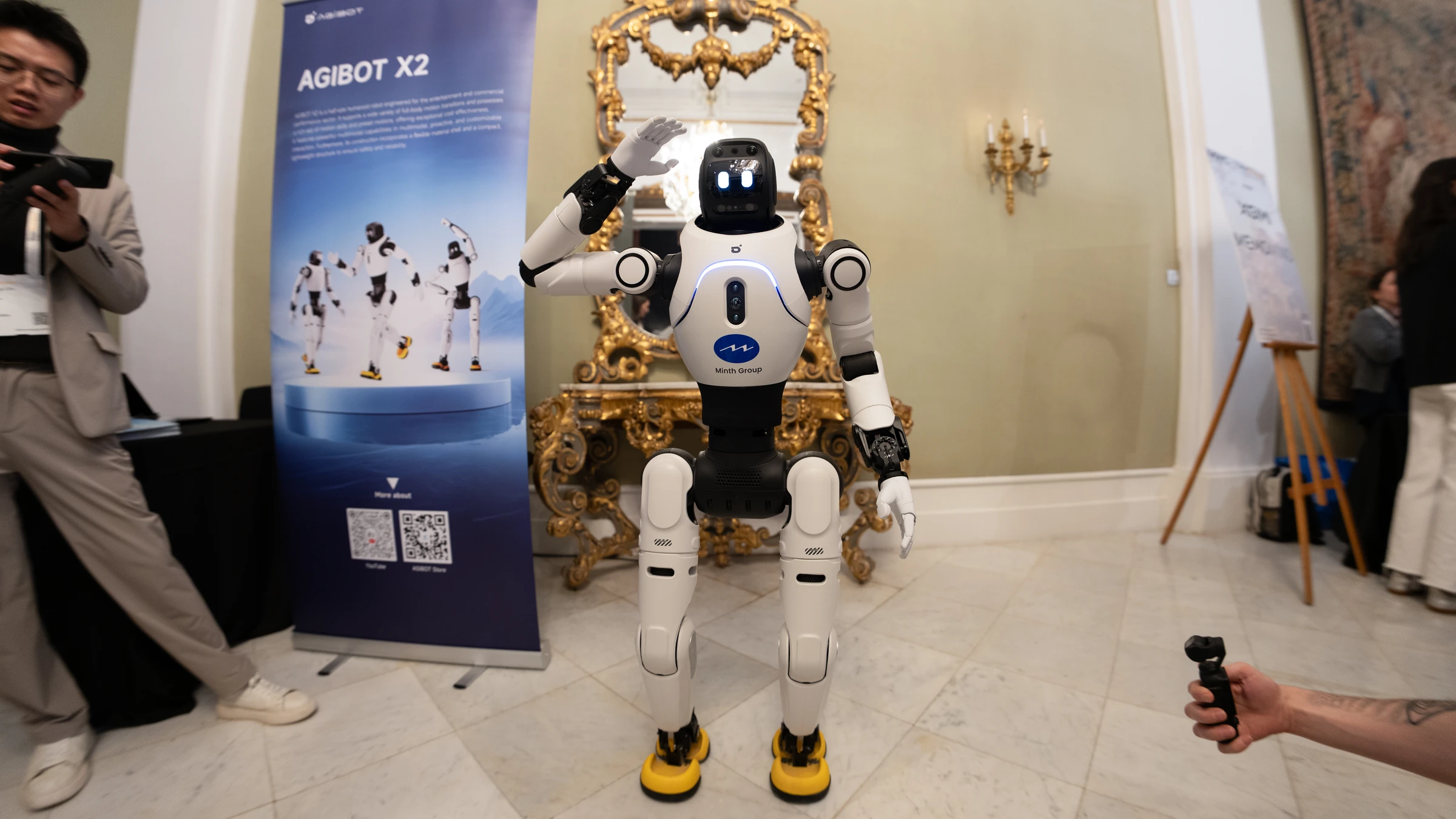 A white AGIBOT X2 humanoid raises one hand beside an AGIBOT X2 sign at MWC Barcelona in March 2026.