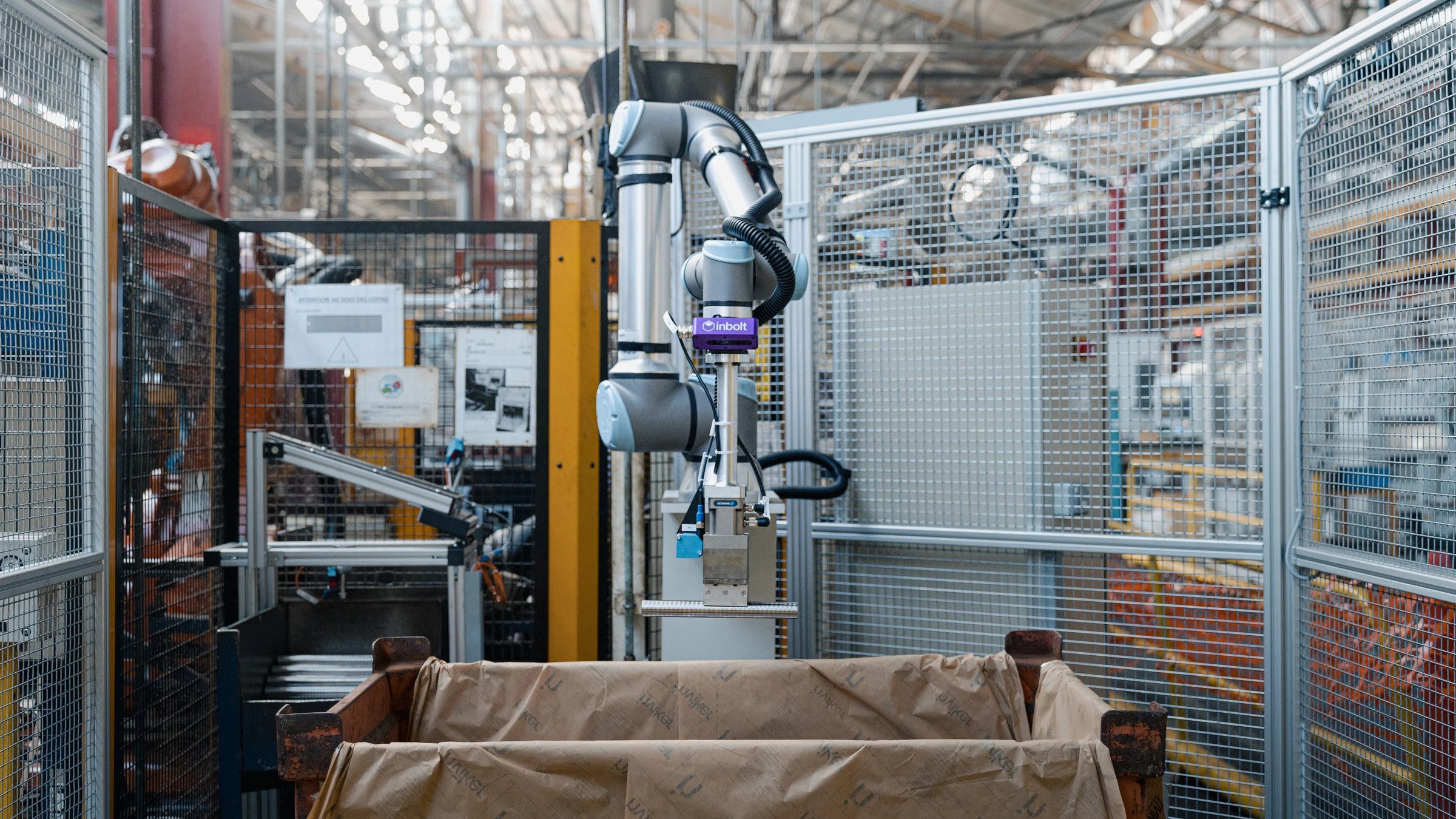 A robot arm with an Inbolt camera above a lined bin inside a fenced factory cell. Contextual company photograph from September 2025.