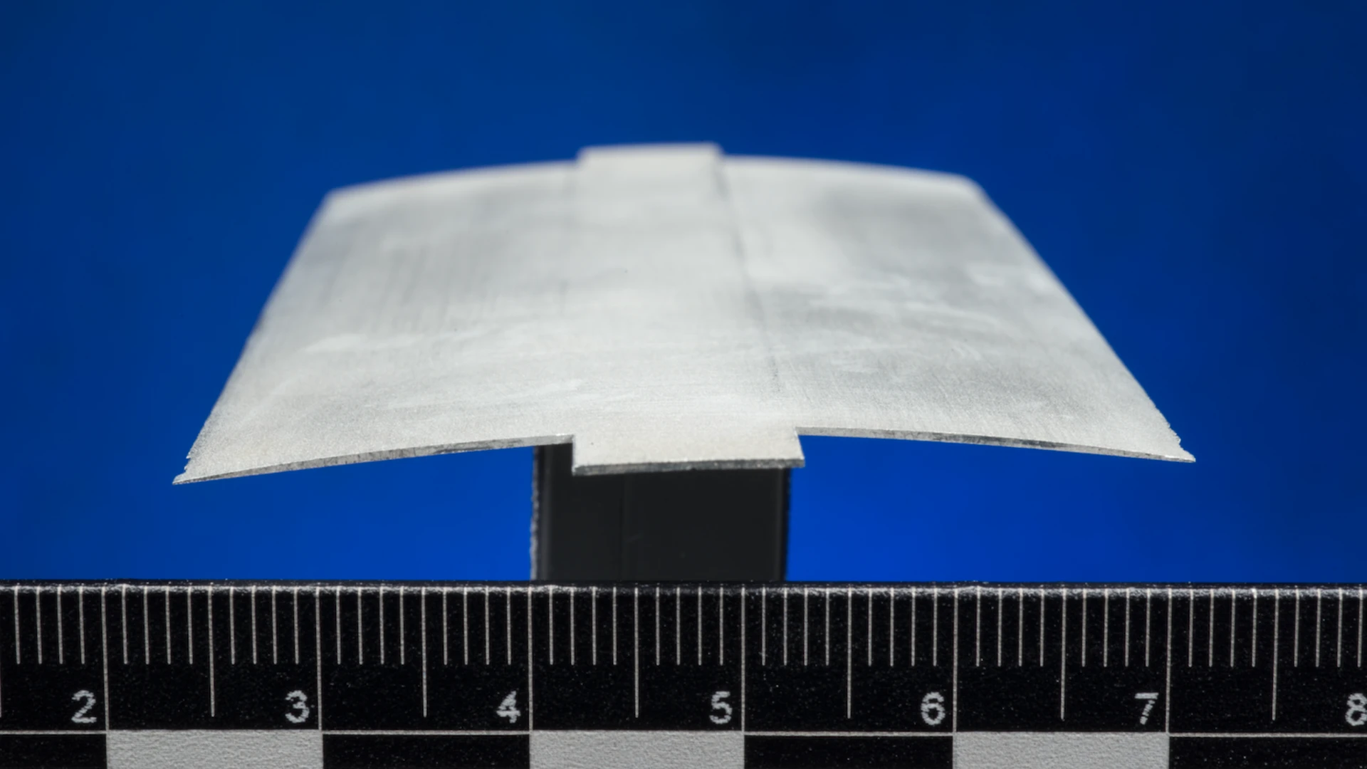 A thin roamx-0201 experimental airfoil sample on a black stand above a ruler, photographed against a blue background.