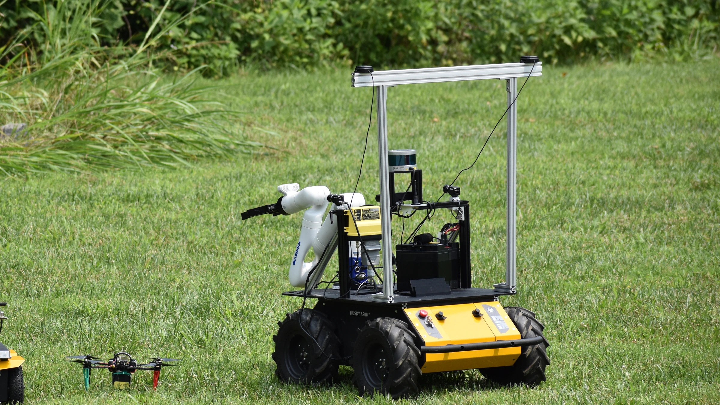 A yellow sampling rover with a white robotic arm stands on grass, with a small drone to its left, during the ASTRA field campaign.