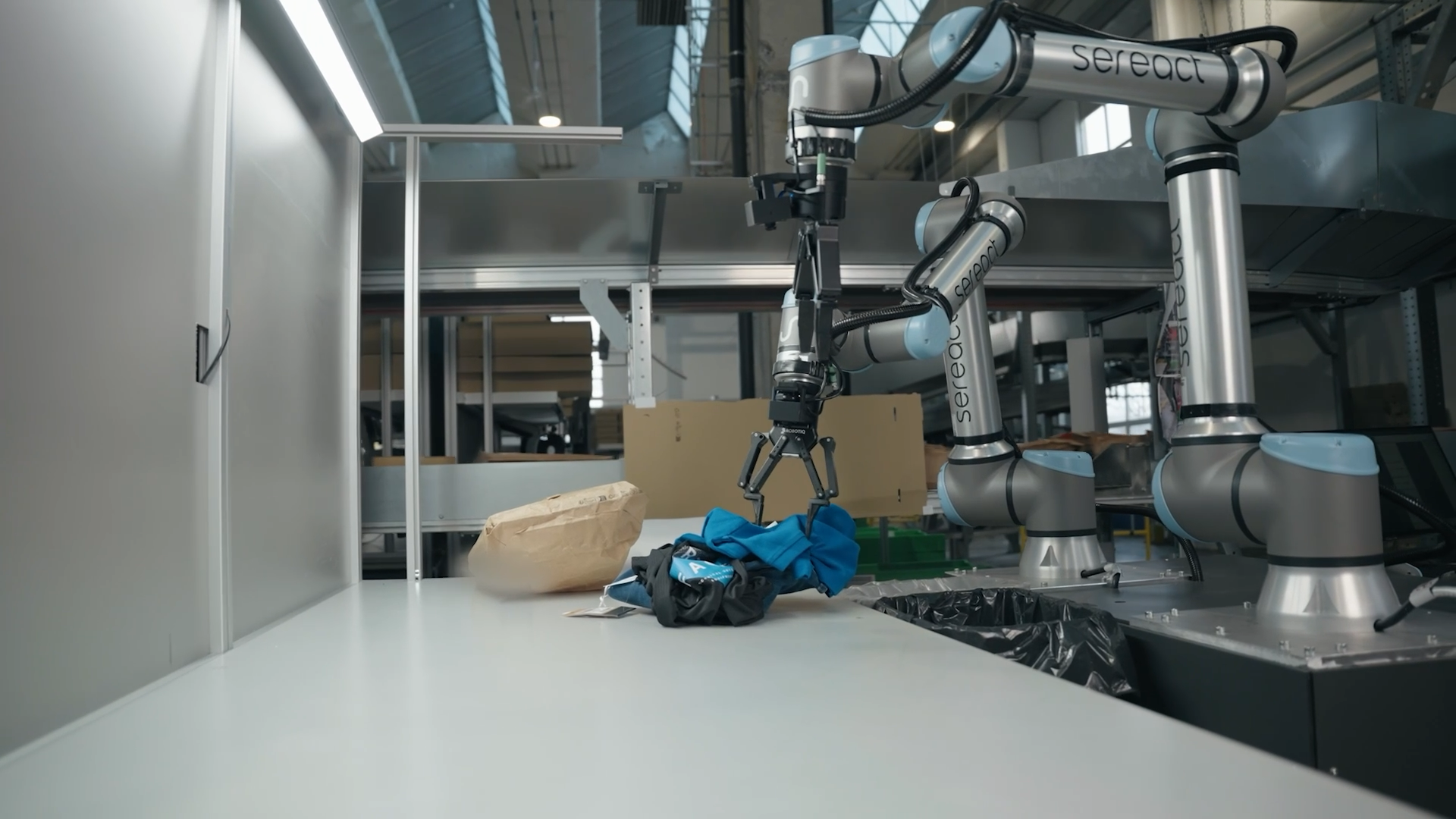 Two Sereact-branded Universal Robots arms handle garments above a work surface in a returns-handling cell.