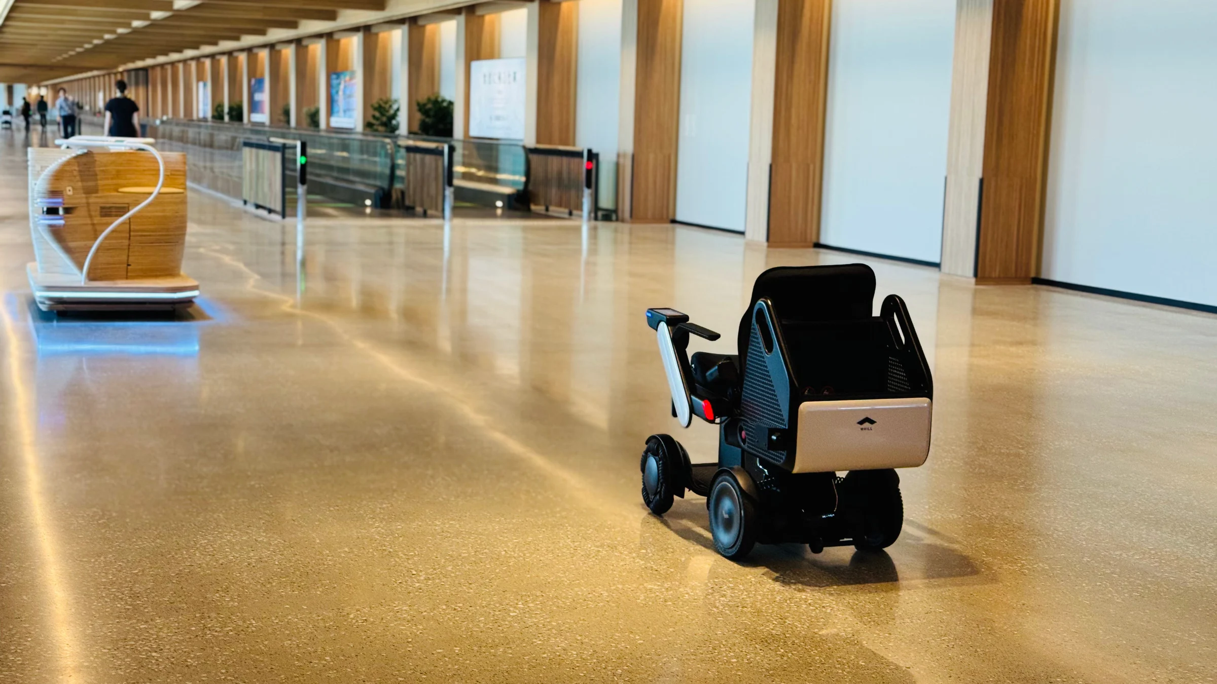 A WHILL autonomous mobility device travels through a Haneda Airport terminal corridor with another autonomous vehicle in the background.